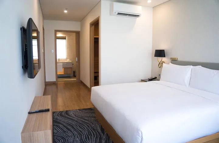 A white bedroom with a double bed, a wall-mounted screen and a small hallway.