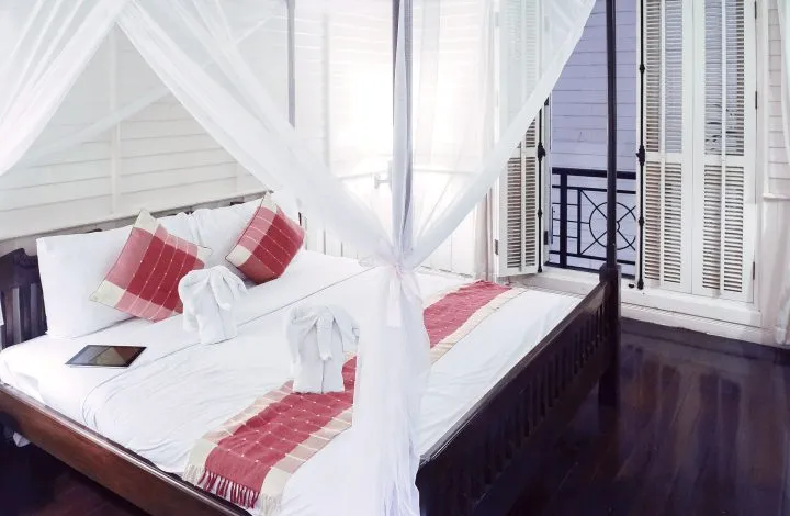 A four-poster bed with white netting and red cushions by a balcony door.