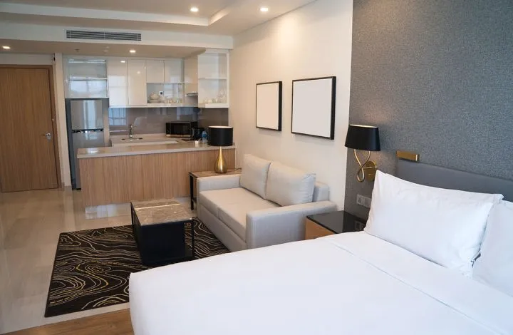 A bedroom with a king bed, a lounge chair and a kitchenette behind.